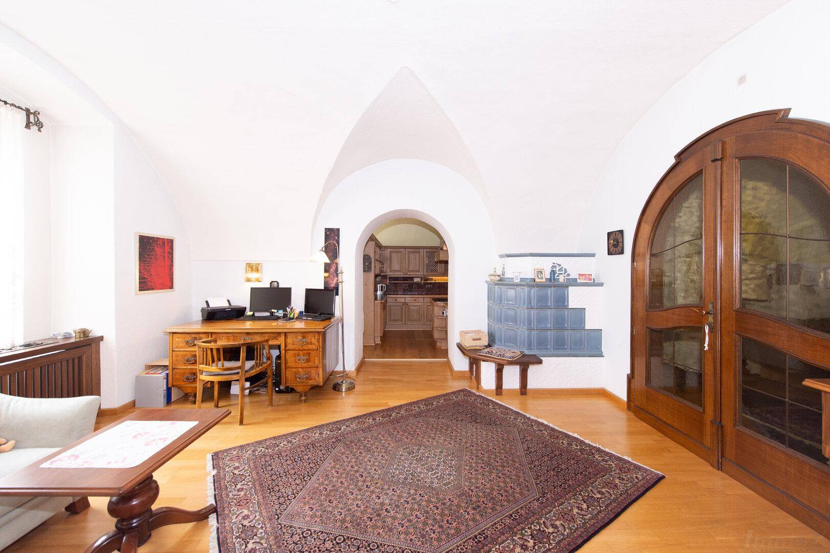 Wohn- und Arbeitsbereich mit gewölbter Decke, Holzboden, Schreibtisch und einem traditionellen Kachelofen.