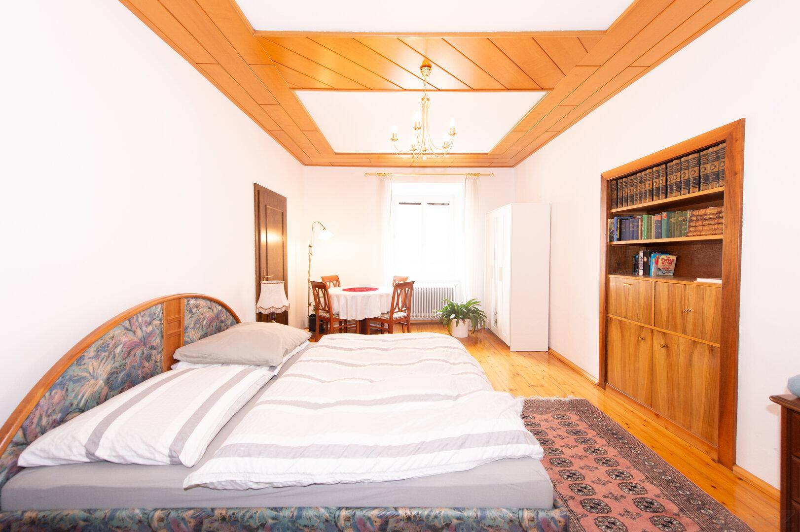 Schlafzimmer mit Bett, Holzdecke, Bücherregal und einem kleinen Essbereich am Fenster.