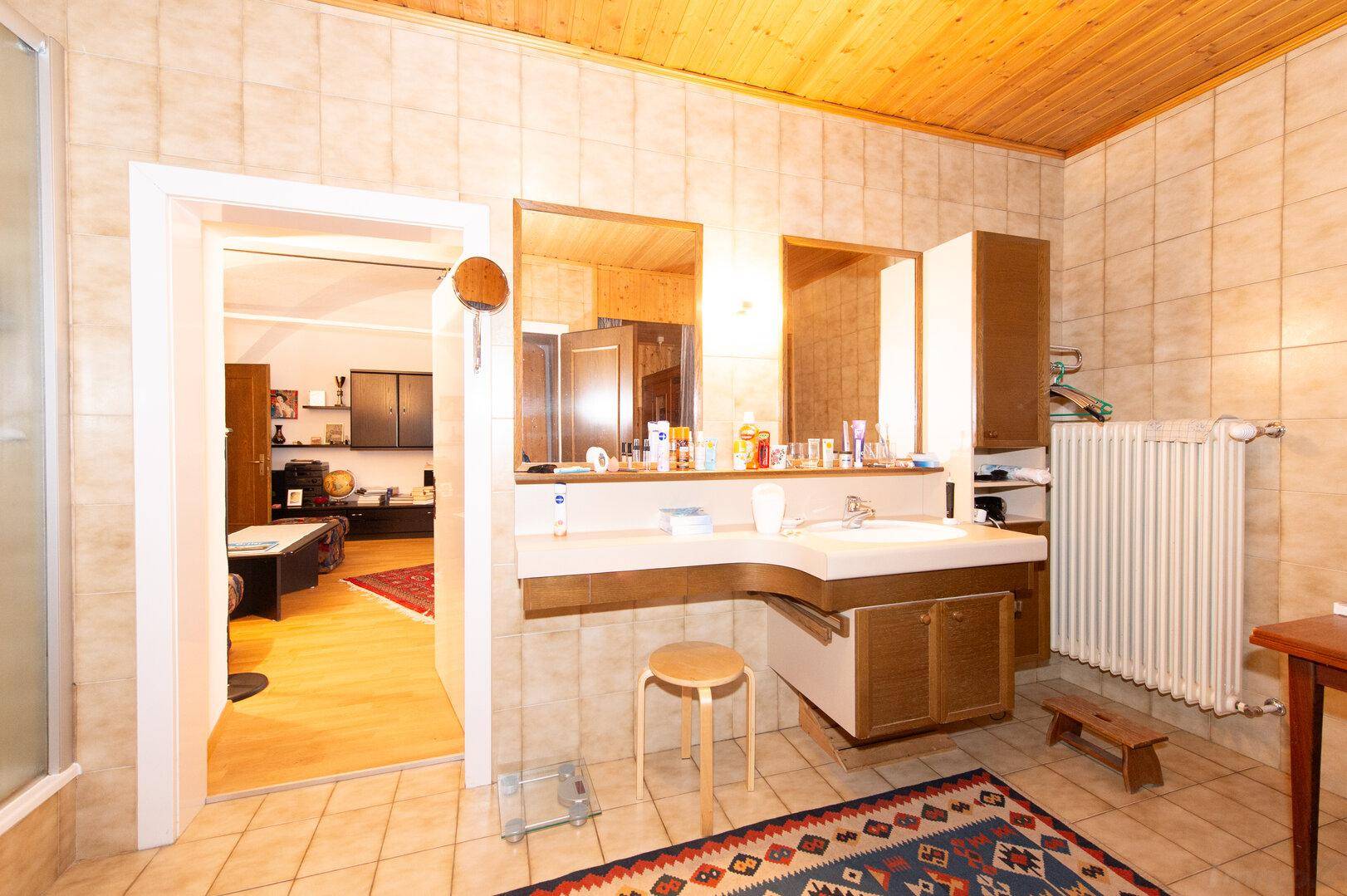 Badezimmer mit Waschtisch, Spiegel, beigefarbenen Fliesen und einer Holzdecke, offen zu einem weiteren Raum.