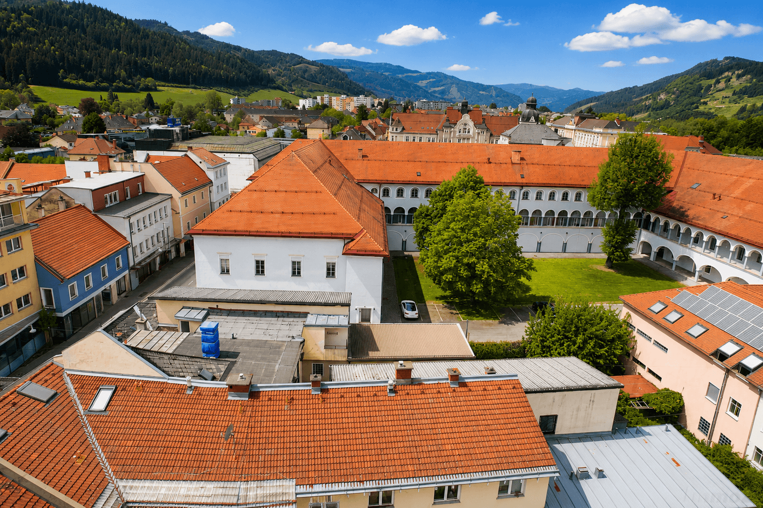 Luftaufnahme einer Stadt mit roten Dächern, umgeben von grünen Hügeln und Bergen.