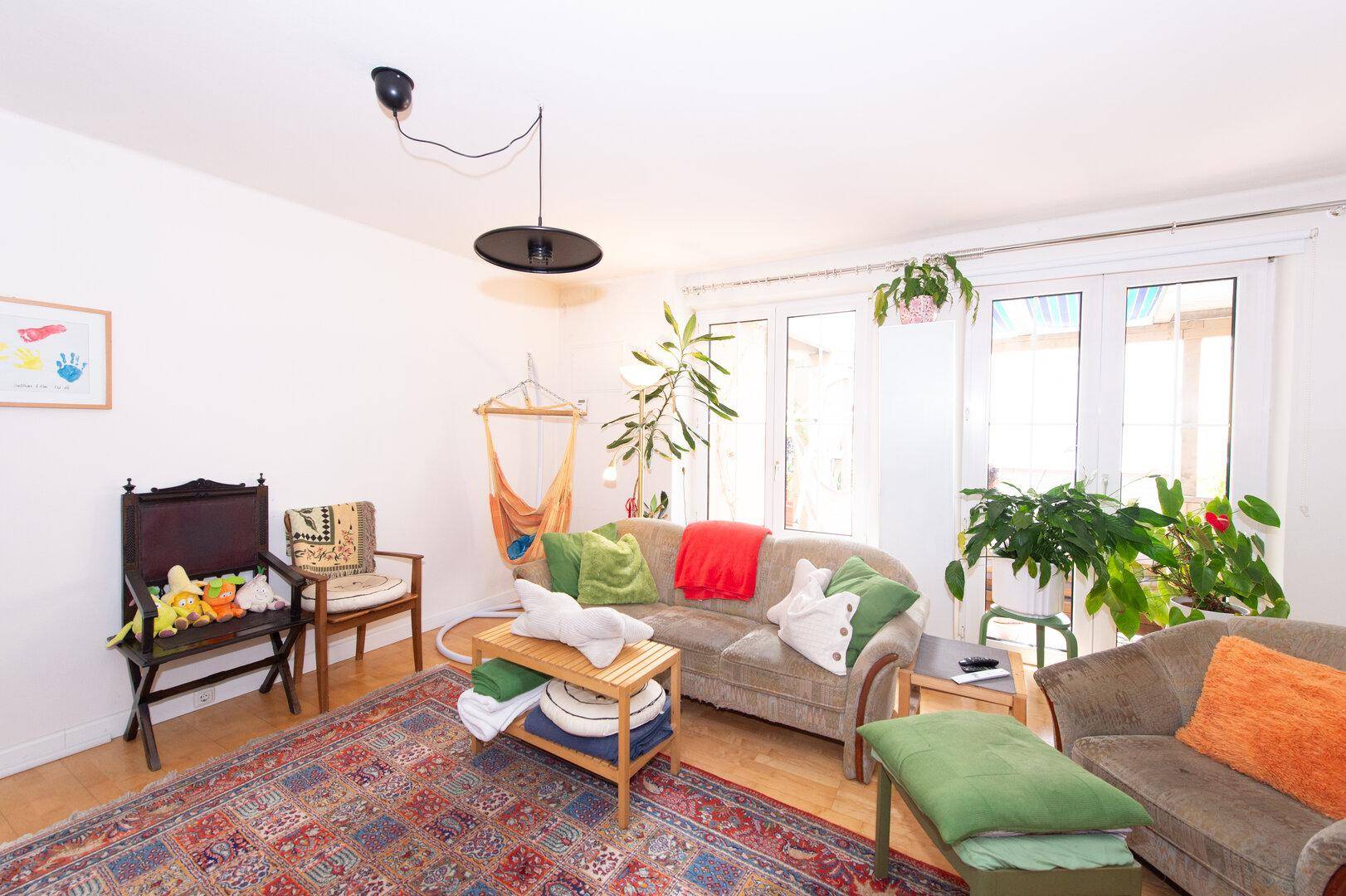 Helles Wohnzimmer mit Sofa, Pflanzen, Hängesessel und großen Fenstern.