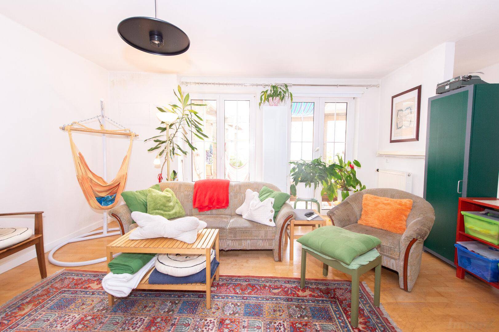 Wohnzimmer mit Sofa, Pflanzen und einem grünen Schrank, hell beleuchtet durch große Fenster.