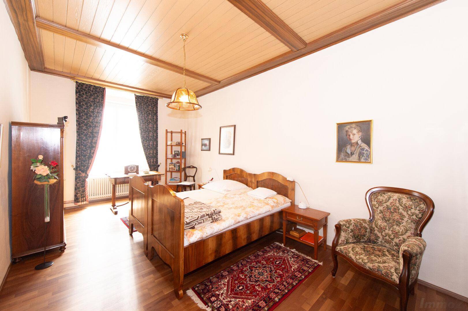 Schlafzimmer mit Holzbett, Holzdeckenbalken und einem Fenster mit dunklen Vorhängen.