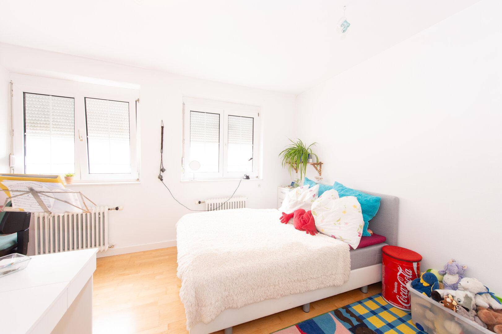 Helles Schlafzimmer mit Holzboden, weißen Wänden und zwei Fenstern mit Rollläden.