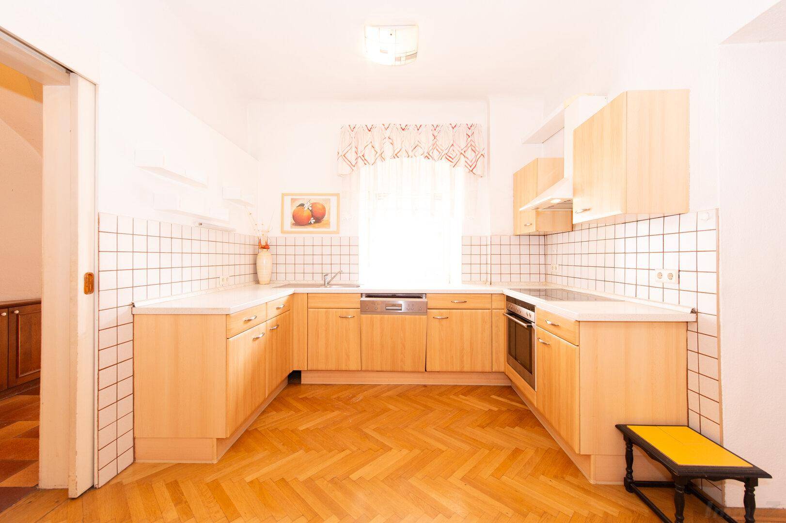Helle Küche mit Holzschränken, weißen Arbeitsplatten und Fischgrätparkettboden.