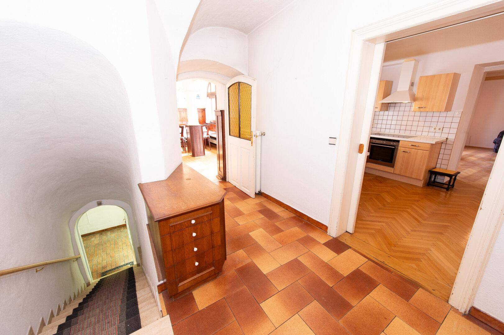 Flur mit Terrakotta-Fliesenboden, Holzkommode und Blick in die Küche sowie ins Treppenhaus.