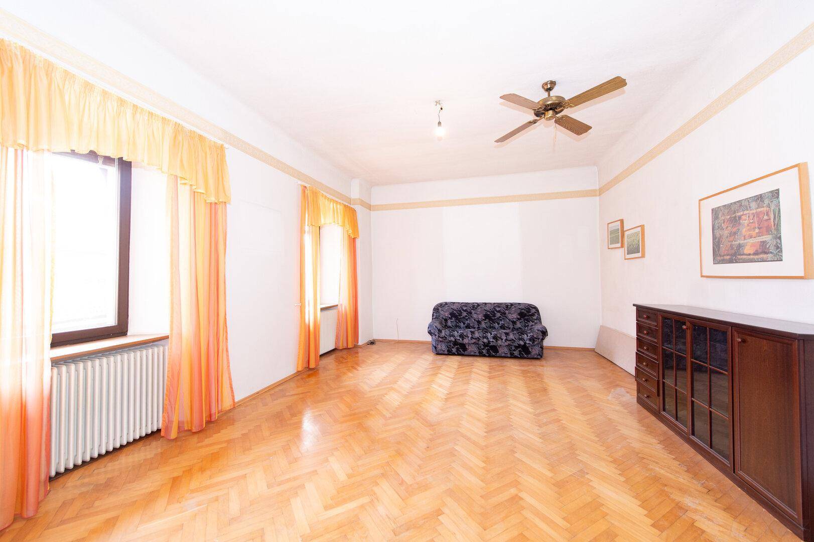 Helles Wohnzimmer mit Fischgrätparkett, großem Fenster und einem dunklen Sofa.