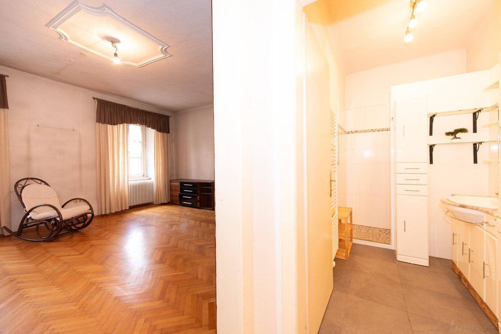 Schlafzimmer mit Parkettboden und Schaukelstuhl, angrenzend an ein Badezimmer mit Dusche.