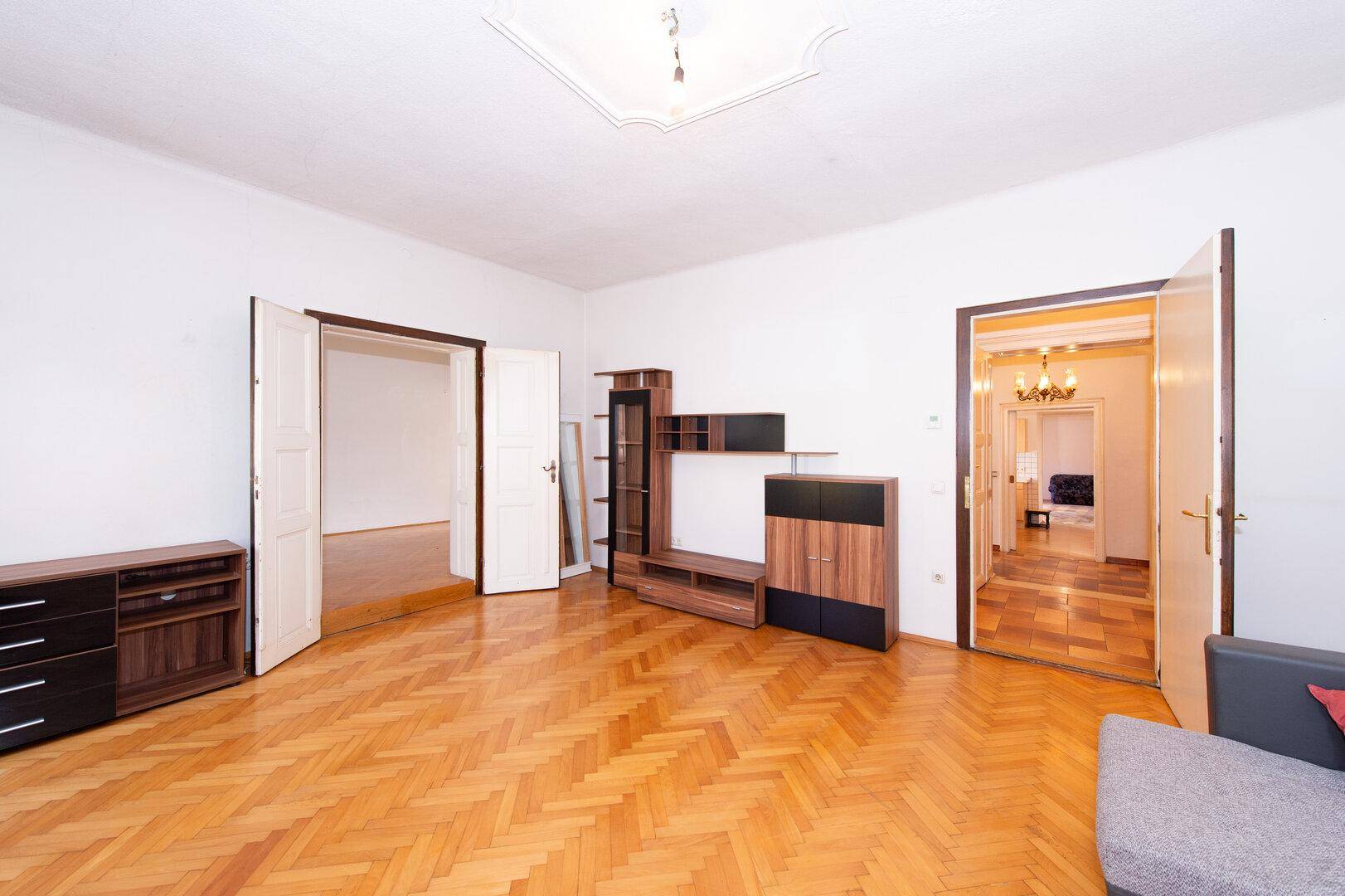 Großes Wohnzimmer mit Fischgrätparkett, dekorativer Decke und Zugang zu weiteren Räumen.
