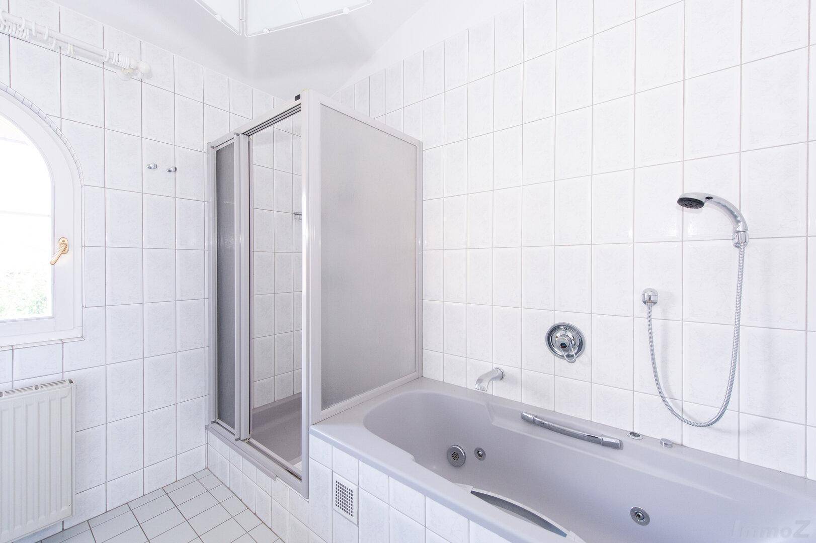 Weiß gefliestes Badezimmer mit Badewanne, Duschkabine und Fenster.