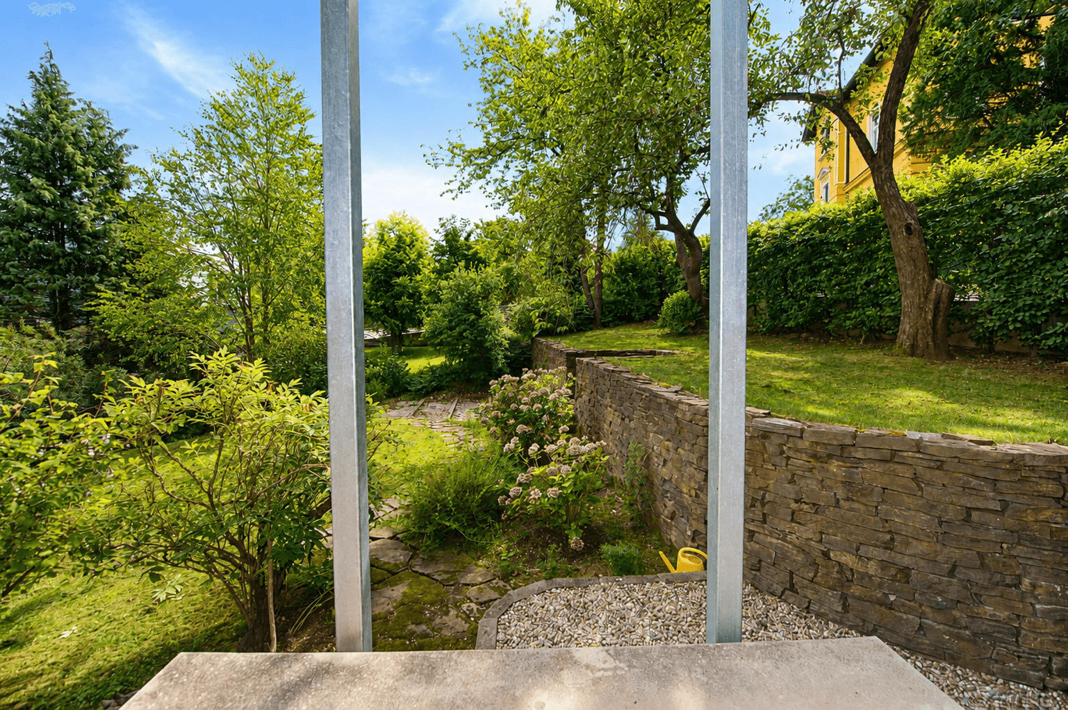 Weitläufiger Gartenbereich mit altem Baumbestand und Steinmauern unter blauem Himmel.
