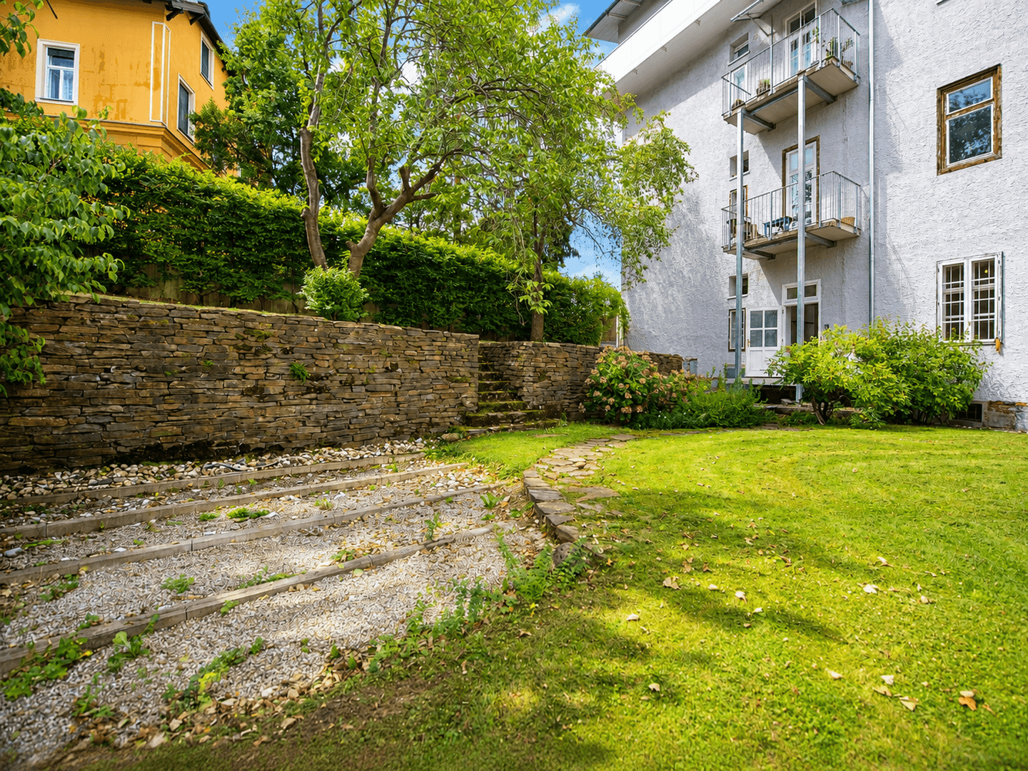 Grüner Gartenbereich mit alter Steinmauer, Kieswegen und Blick auf die Gebäudefassade.