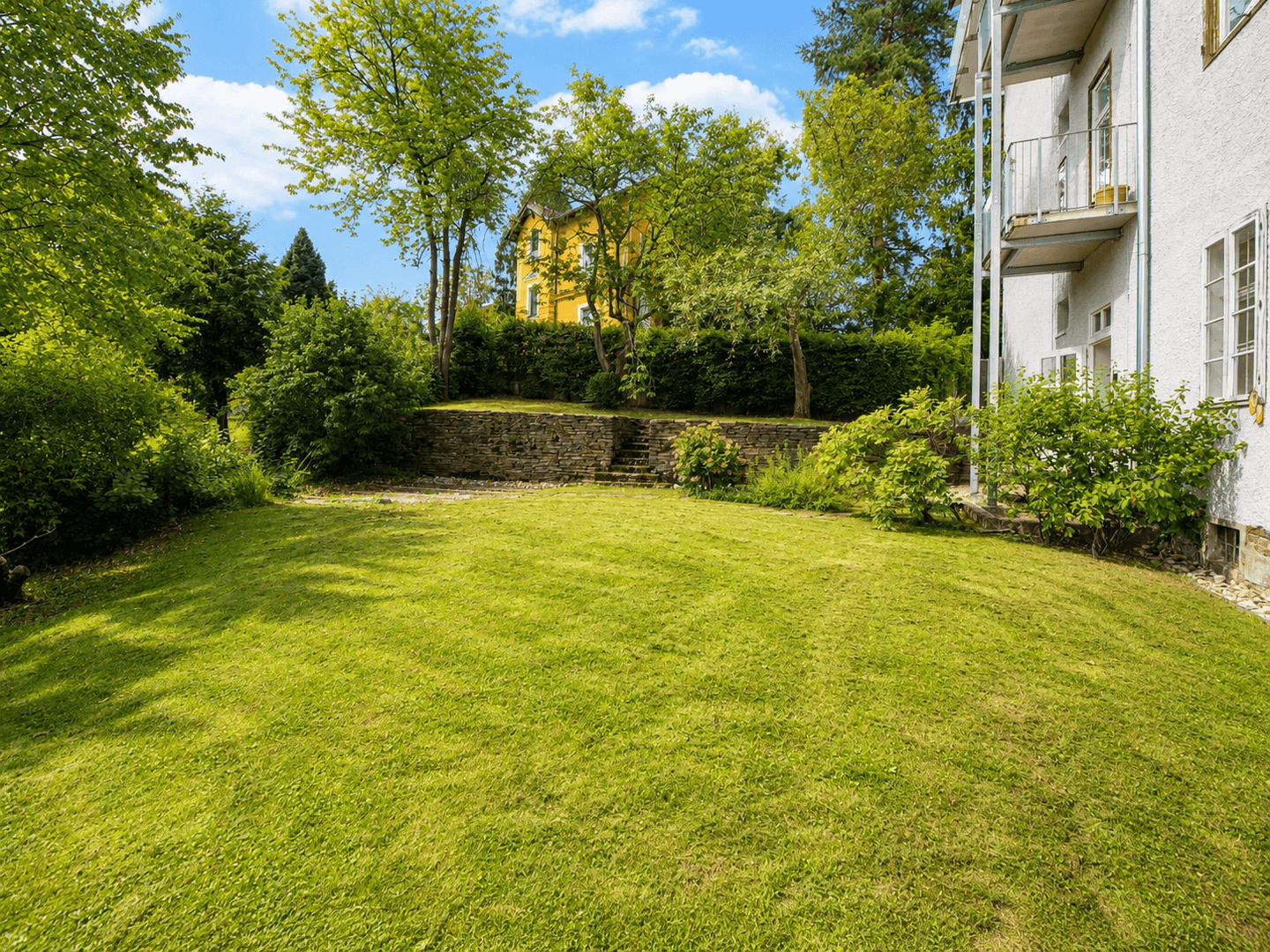 Große Rasenfläche im Garten mit Hecken und einem gelben Nachbargebäude im Hintergrund.