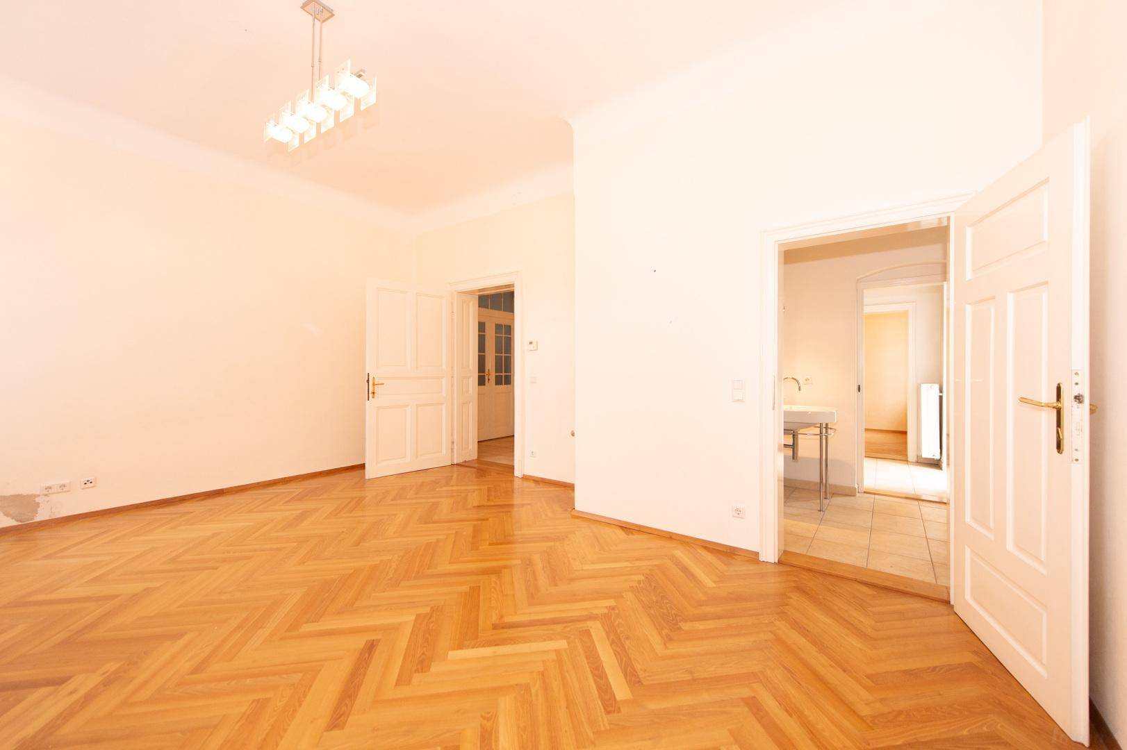 Geräumiger Wohnraum mit Fischgrätparkett und Zugang zu weiteren Zimmern der Wohnung.