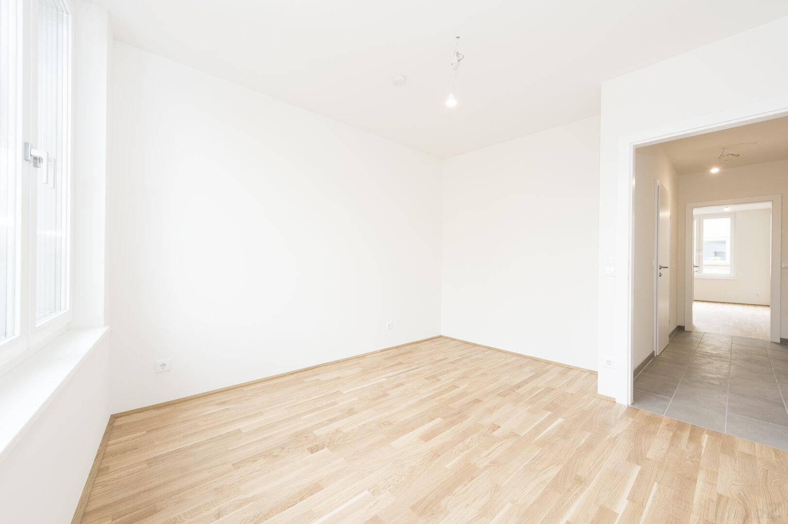 Leeres Zimmer mit Holzboden, großem Fenster und Blick in den gefliesten Flur.