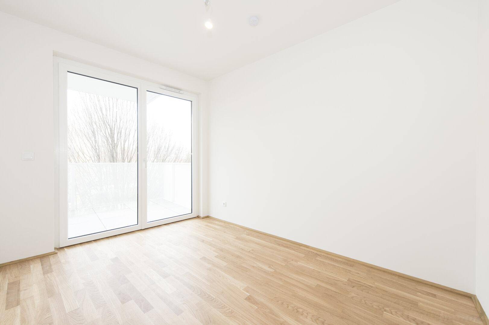 Helles Zimmer mit Holzboden und großer Schiebetür, die auf einen Balkon führt.