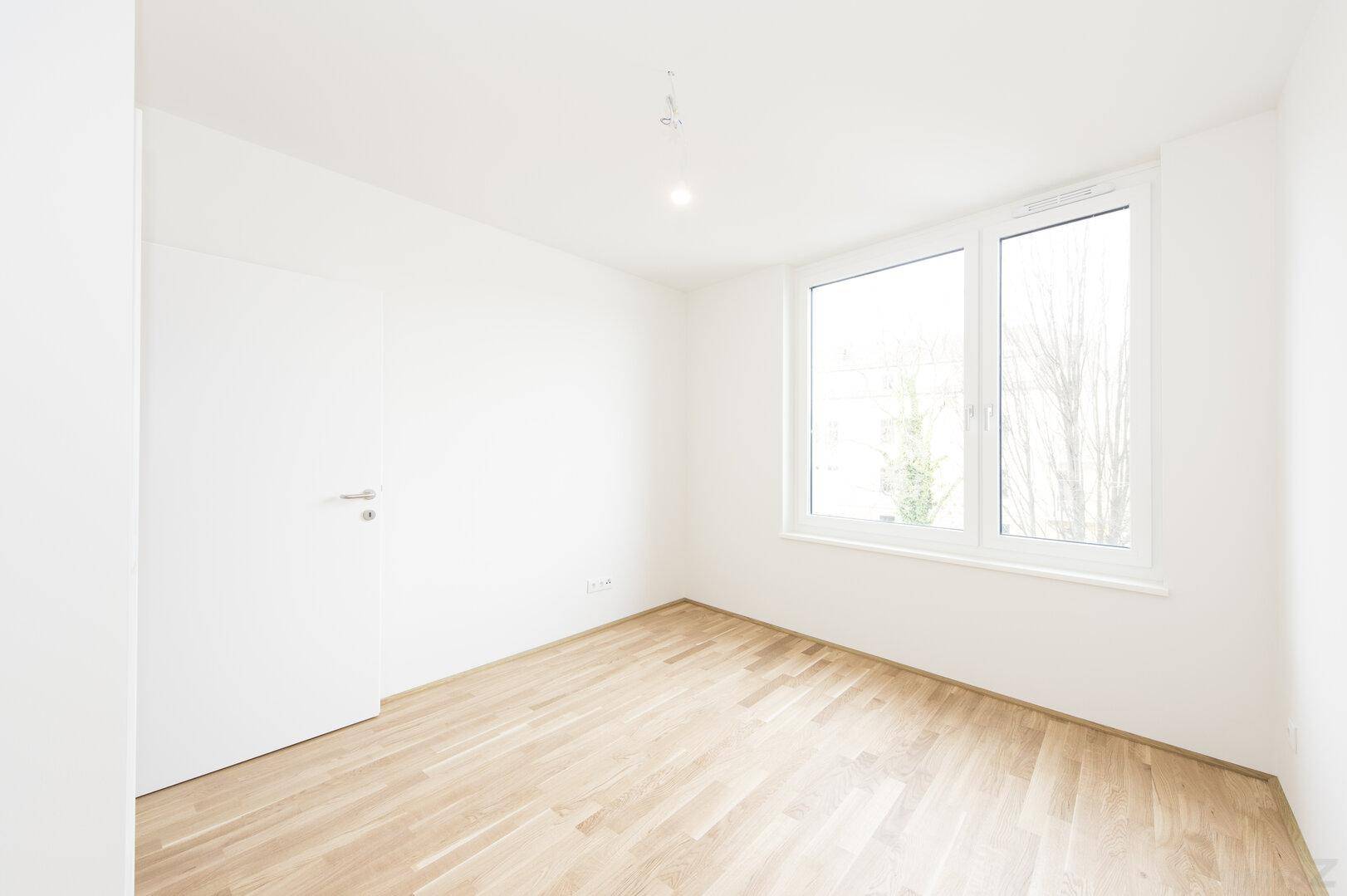 Helles Zimmer mit Holzboden und einem großen Fenster, das viel Tageslicht hereinlässt.