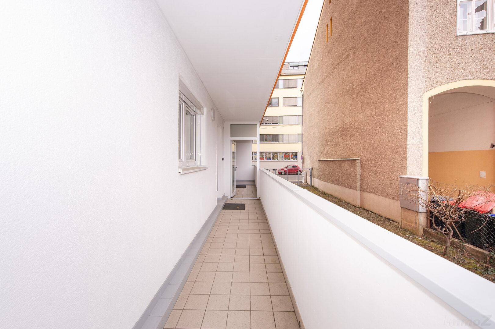 Langer Balkon mit Fliesenboden und Blick auf die umliegenden Gebäude und die Straße.
