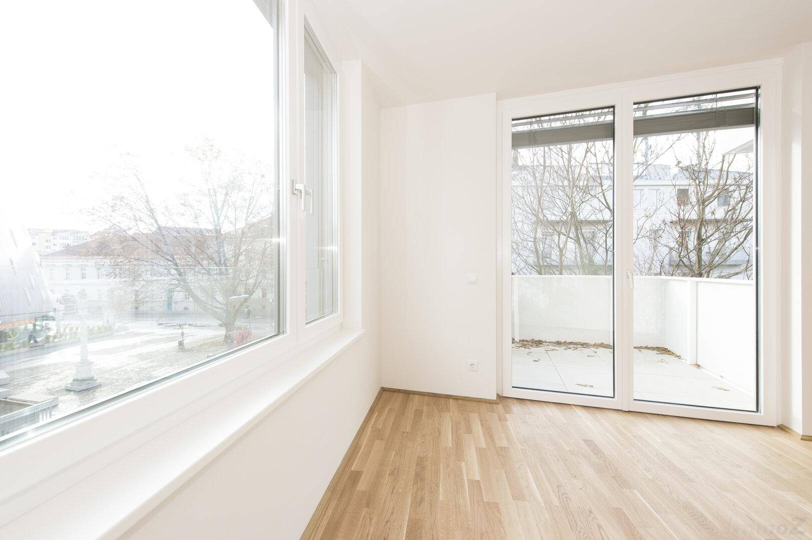 Heller Wohnraum mit großen Fenstern, Parkettboden und Zugang zu einem Balkon.