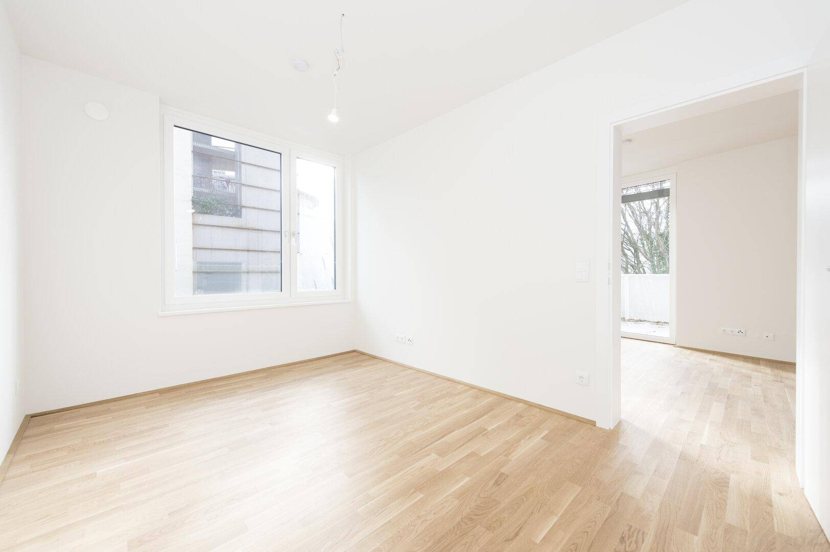 Leeres Zimmer mit hellem Parkettboden und einem großen Fenster, das viel Licht hereinlässt.