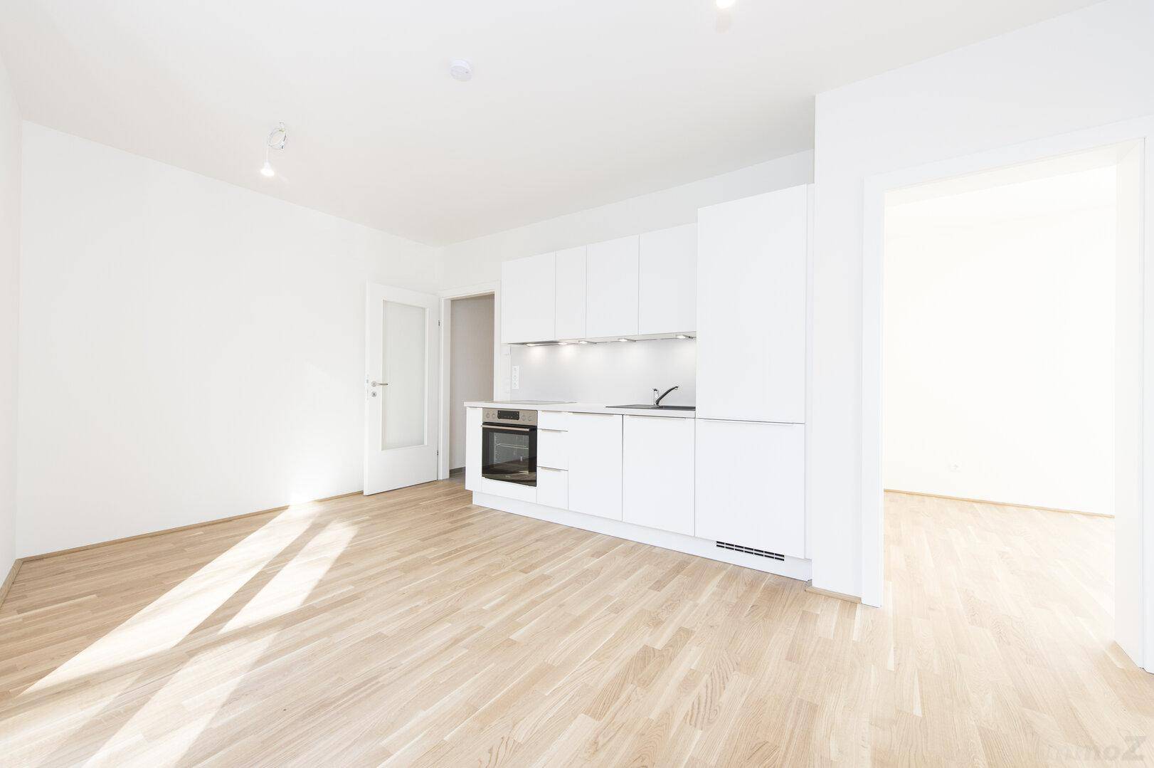 Kompakte, moderne weiße Einbauküche in einem hellen Raum mit Holzfußboden und Zugang zu weiteren Zimmern.