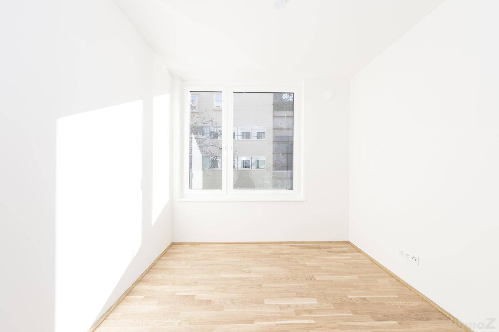 Helles, leeres Schlafzimmer mit Holzfußboden und einem großen Fenster, das viel Sonnenlicht hereinlässt.