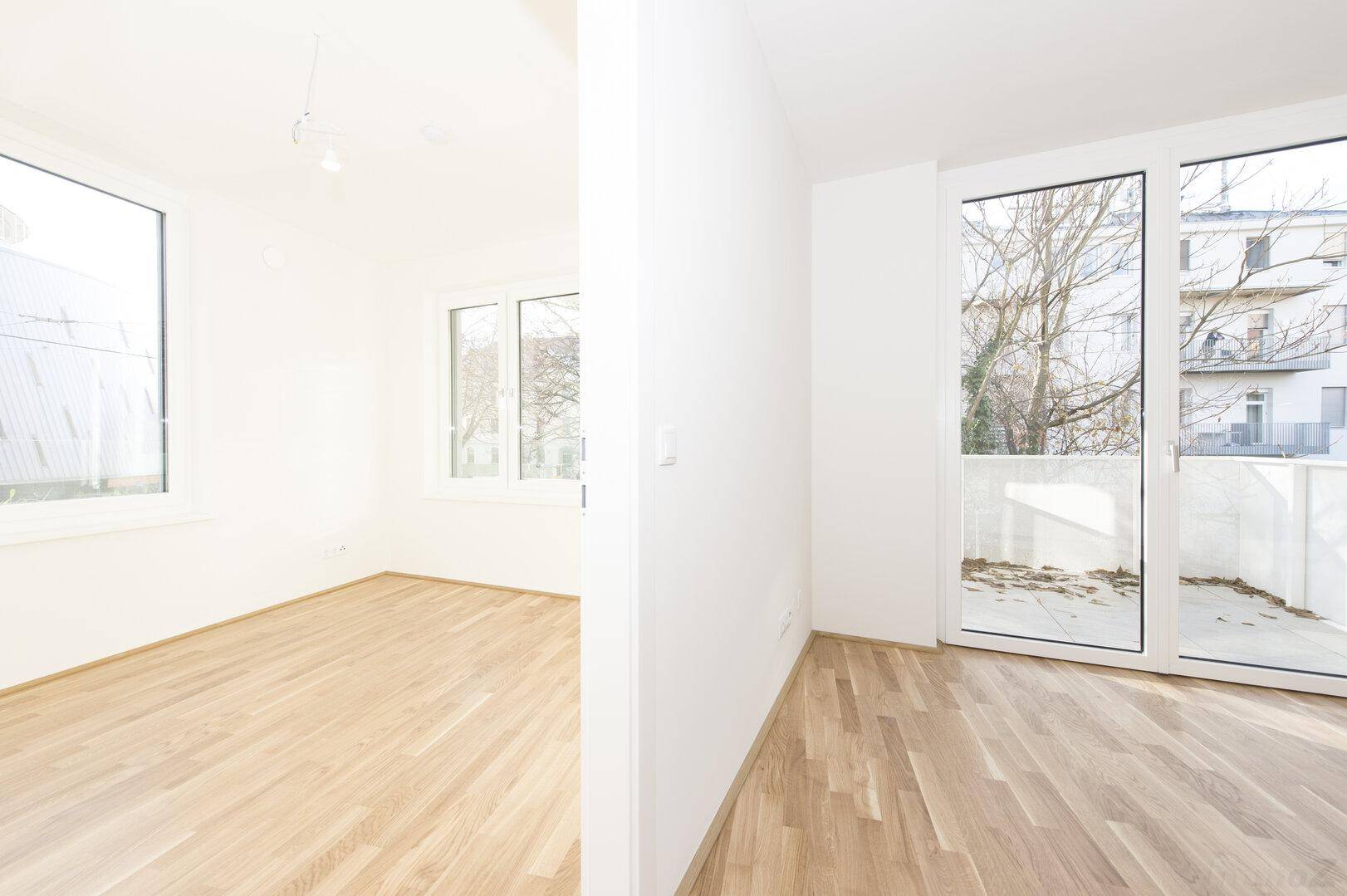 Zwei helle, leere Räume mit Holzfußboden, großen Fenstern und Zugang zu einem Außenbereich.