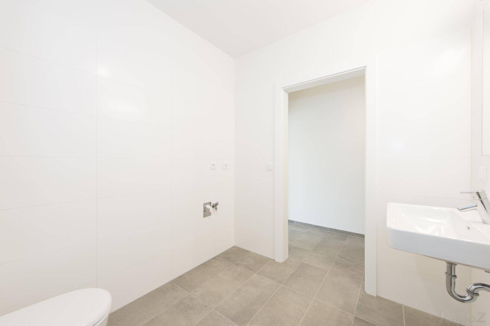 Helles Badezimmer mit grauen Bodenfliesen, weiß gefliesten Wänden, Waschbecken und wandhängendem WC.