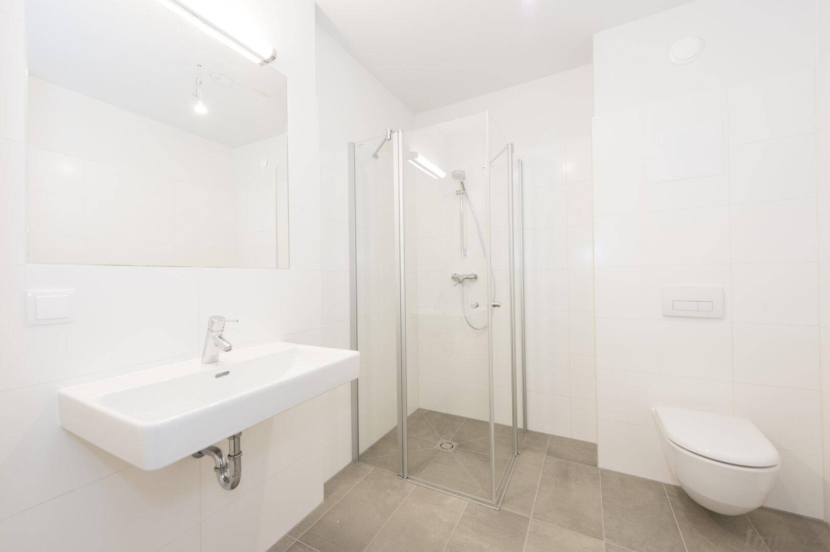 Modernes Badezimmer mit grauen Bodenfliesen, weißer Wandverkleidung, Duschkabine und wandhängendem WC.