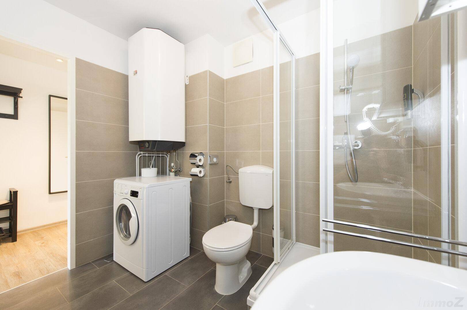 Badezimmer mit Dusche, WC, Waschmaschine und beigefarbenen Fliesen.