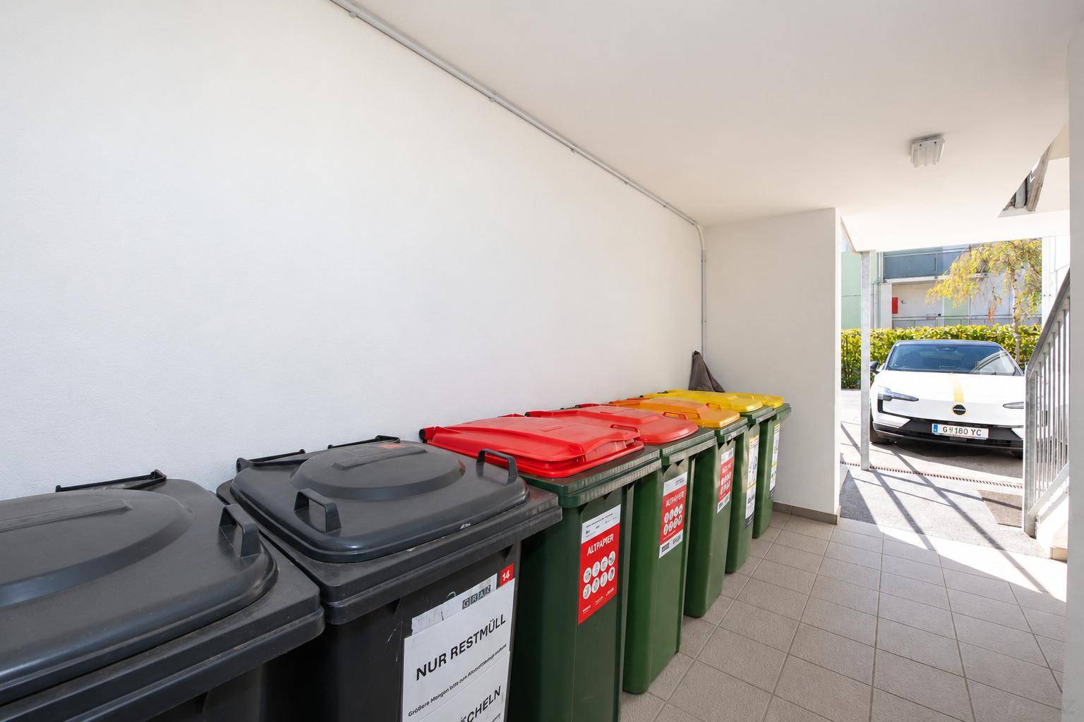 Innenbereich mit mehreren farbigen Mülltonnen und Fliesenboden, ein Auto ist draußen sichtbar.