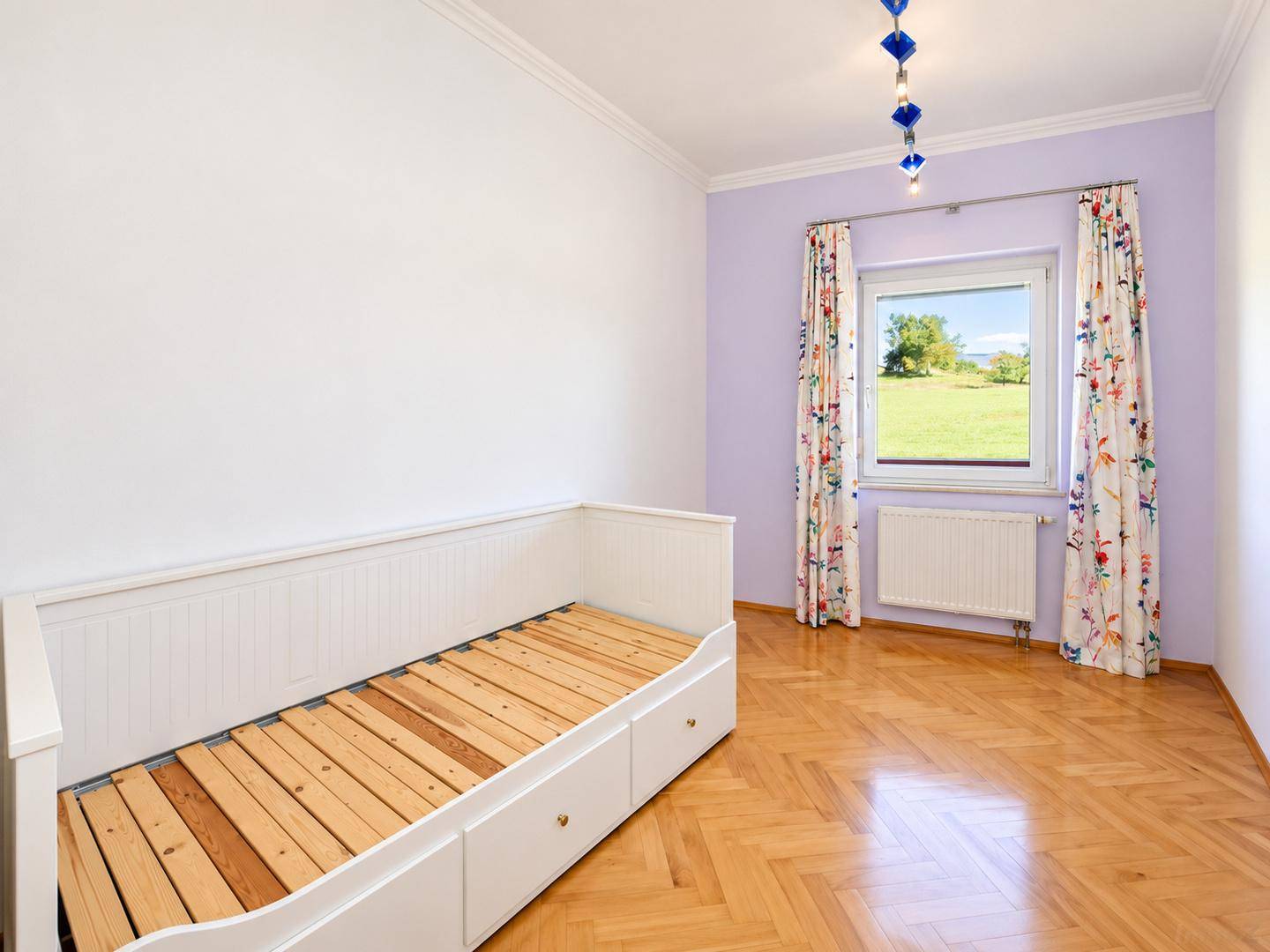 Freundliches Schlafzimmer mit Fischgrätparkett, Tagesbett und farbenfrohen Vorhängen am Fenster.