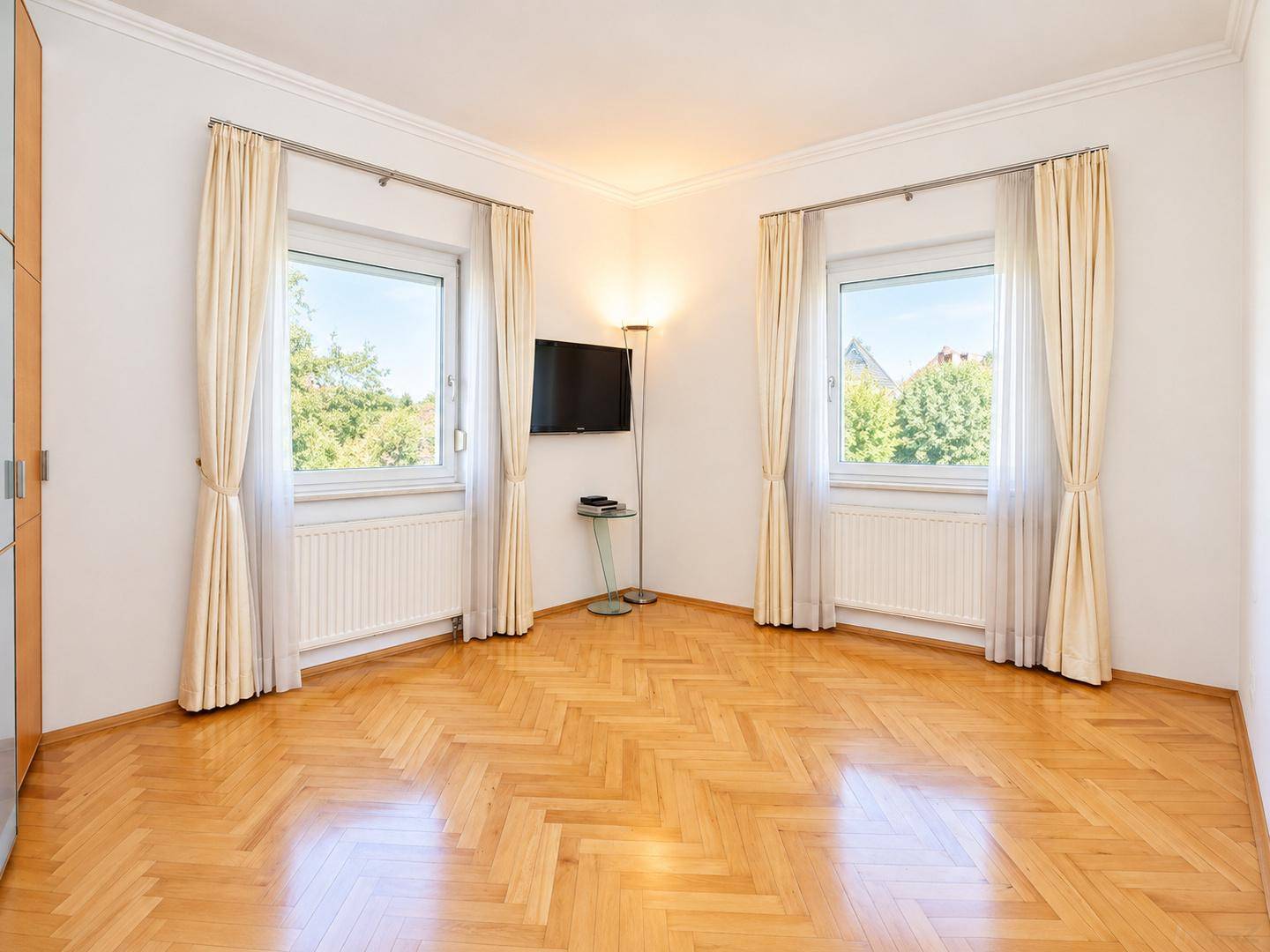 Helles Schlafzimmer mit Fischgrätparkett, zwei Fenstern und einem Wandfernseher.