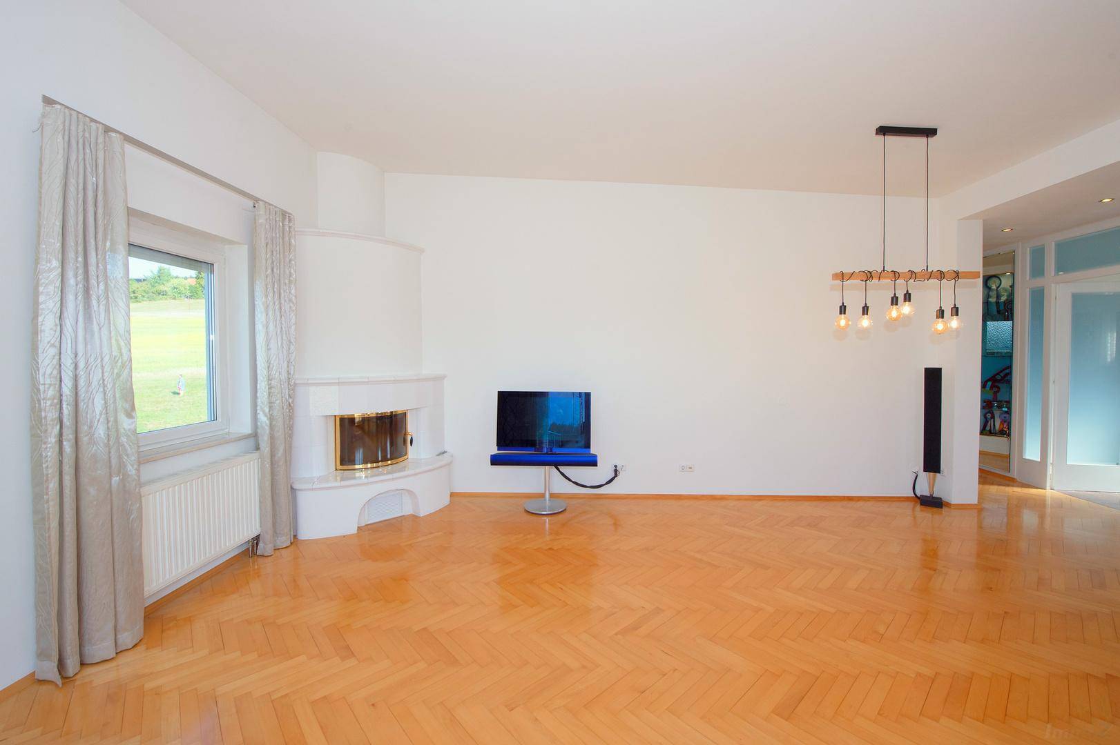 Helles Wohnzimmer mit Kamin, Parkettboden und großem Fenster mit Grünblick.