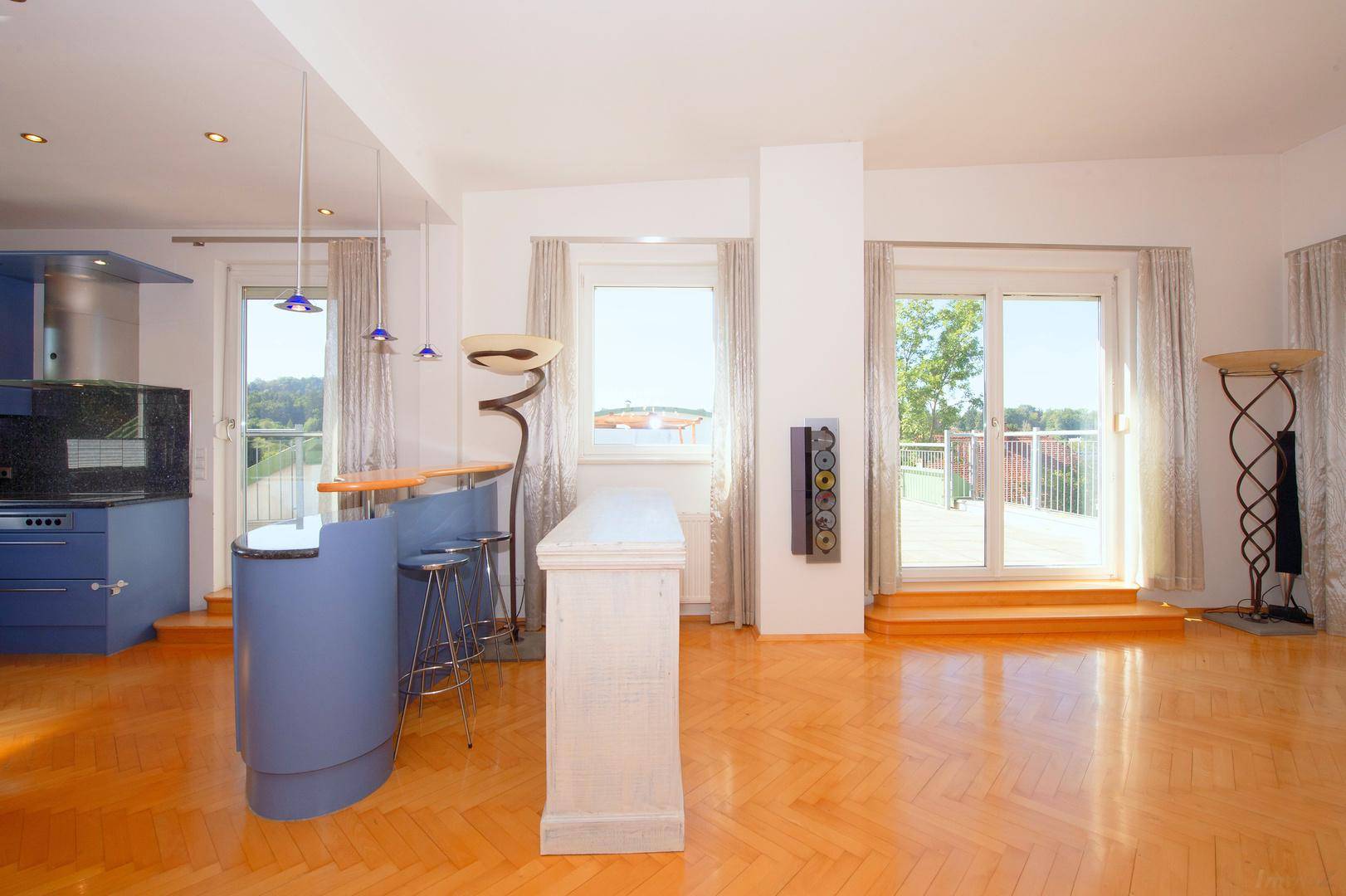 Heller offener Wohnbereich mit blauer Küchenzeile, Bar und Zugang zur Terrasse.