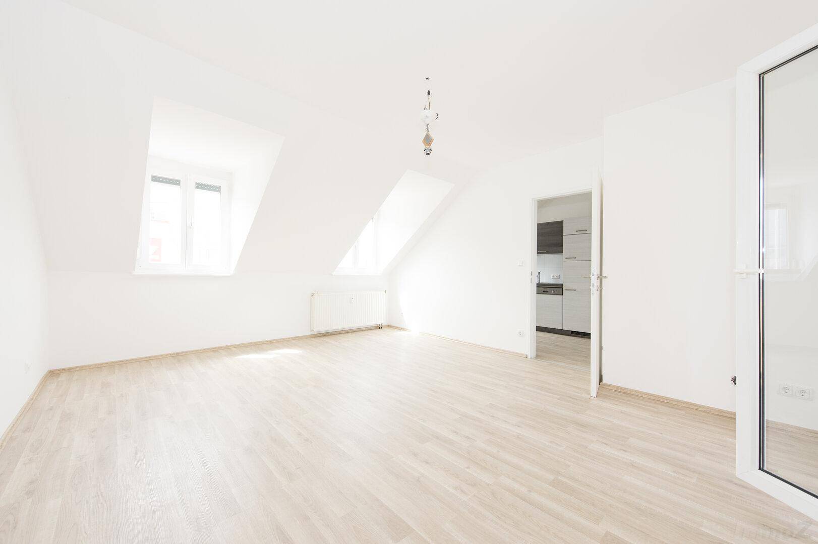 Lichtdurchfluteter Wohnraum mit Dachschrägen, Laminatboden und Blick in die Küche.