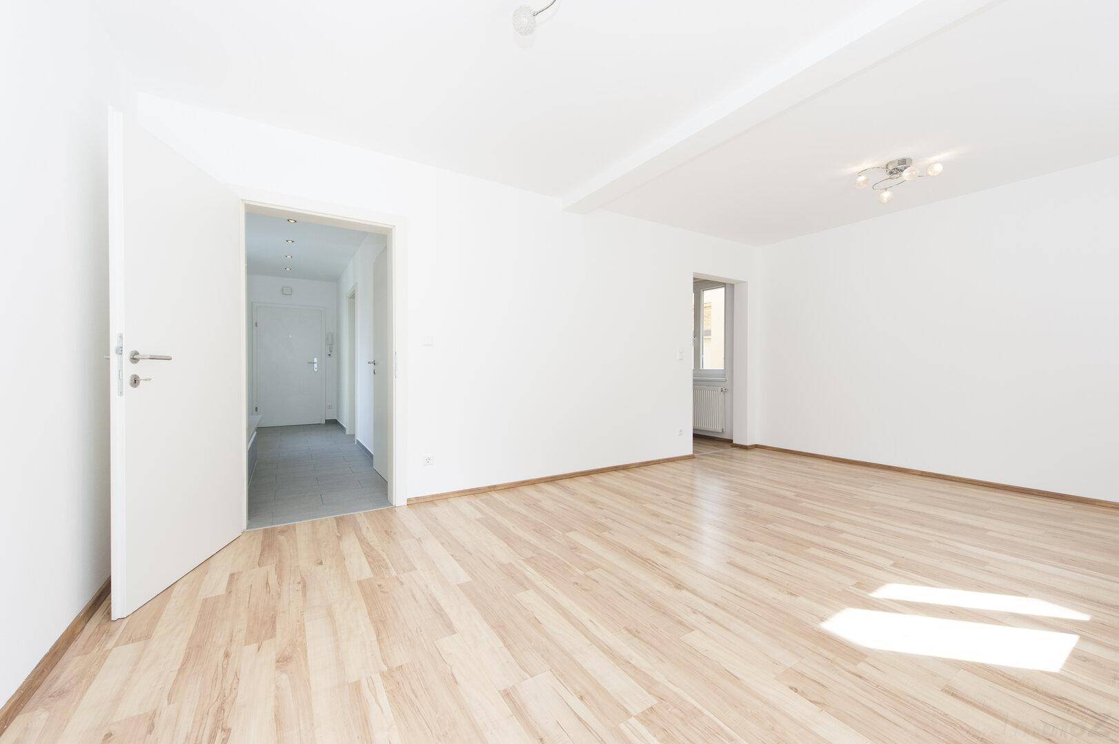 Geräumiges Wohnzimmer mit hellem Laminatboden und Zugang zum Flur und weiteren Räumen.
