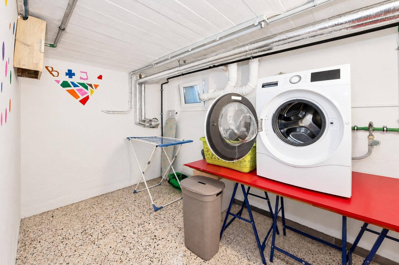 Praktischer Waschraum im Keller mit zwei Waschmaschinen, Trockenständer und sichtbaren Leitungen.