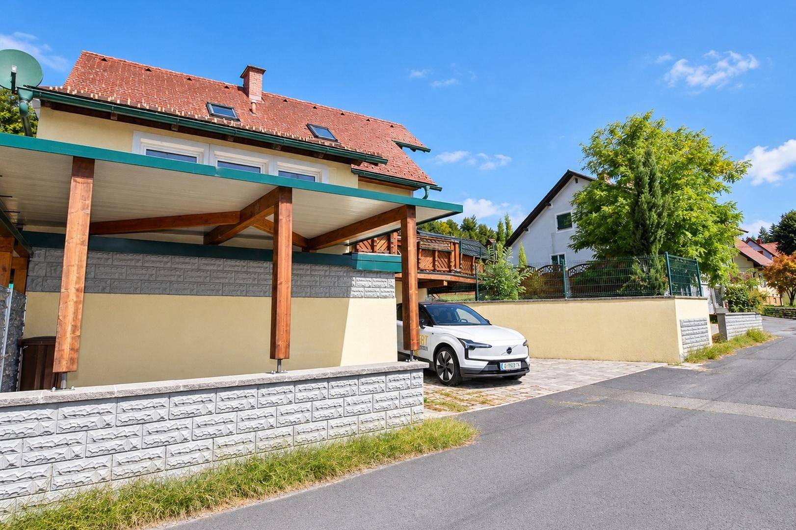 Ansicht des Hauses mit überdachtem Stellplatz und Einfahrt, umgeben von einer Steinmauer und Bäumen.