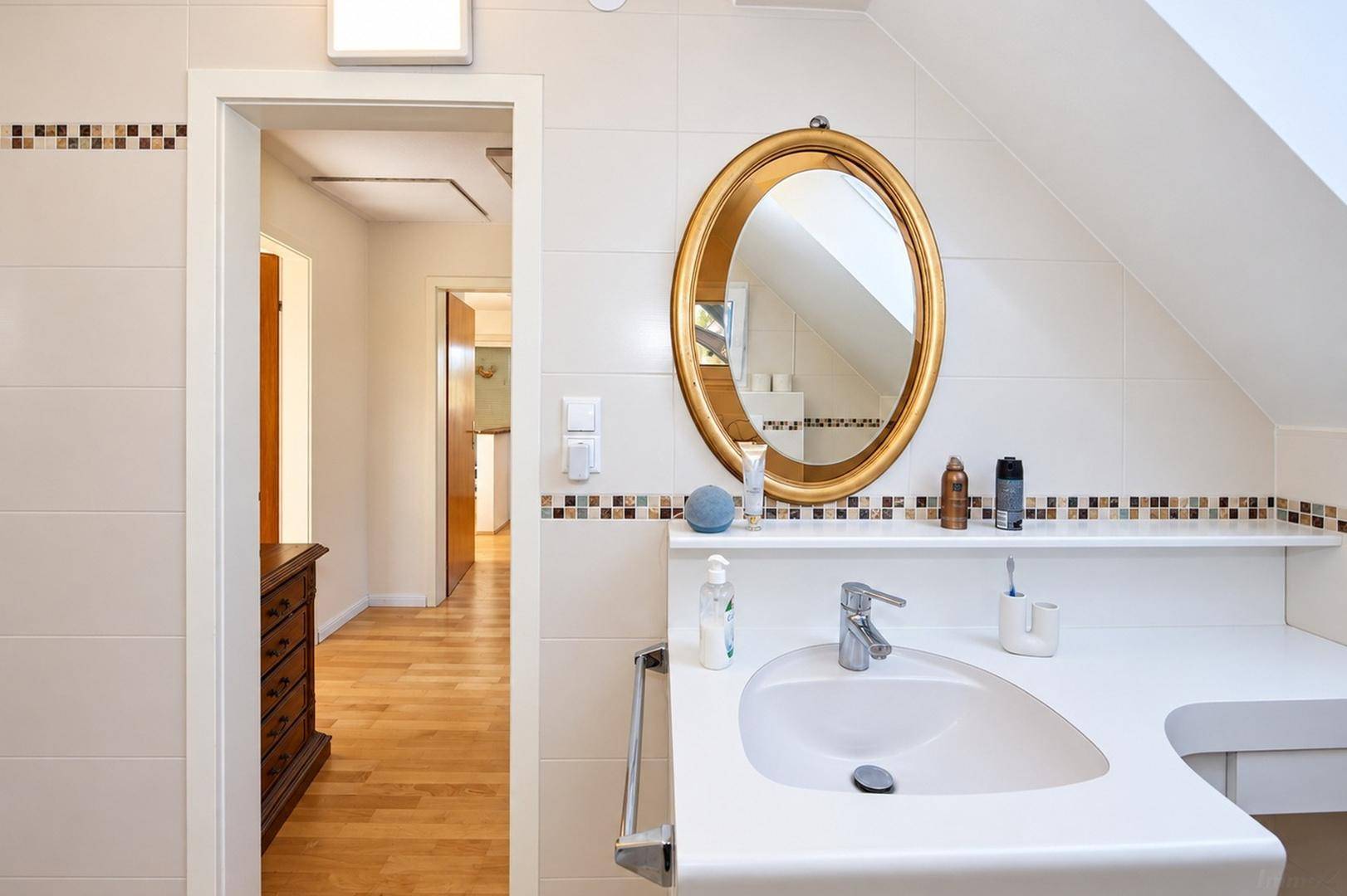 Helles Badezimmer mit Waschbecken, ovalem Spiegel und Dachschräge, gefliest in Weiß.