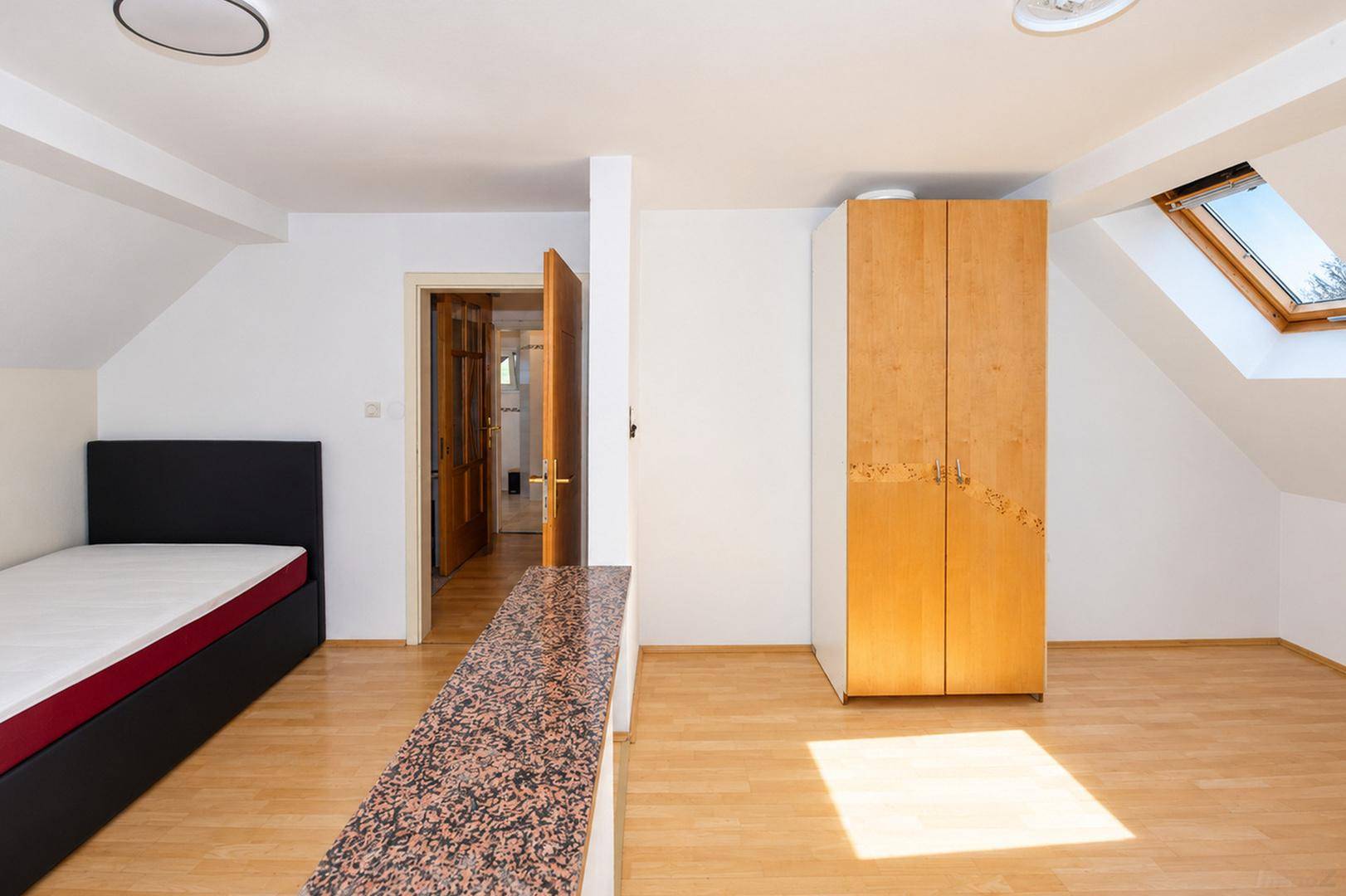 Offener Raum mit Einzelbett, Holzschrank, Holzboden und Dachfenster unter Schrägdach.
