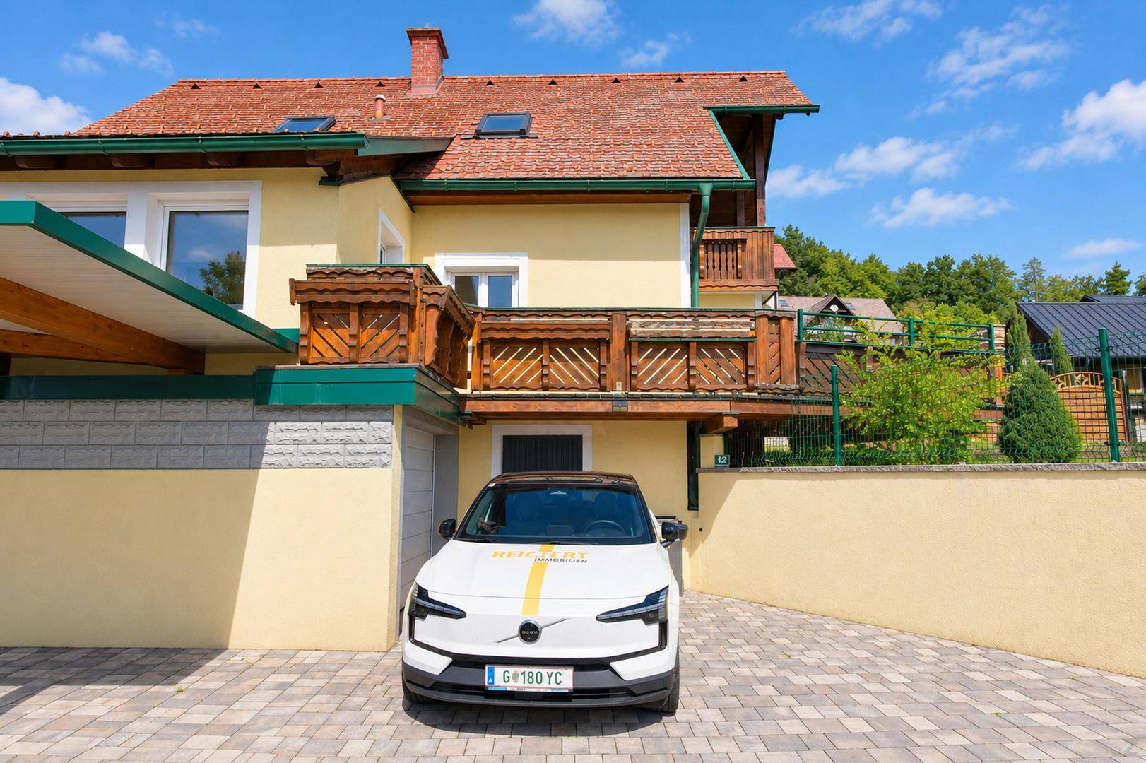 Gelbe Hausfassade mit rotem Ziegeldach und gepflasterter Einfahrt mit parkendem Auto.
