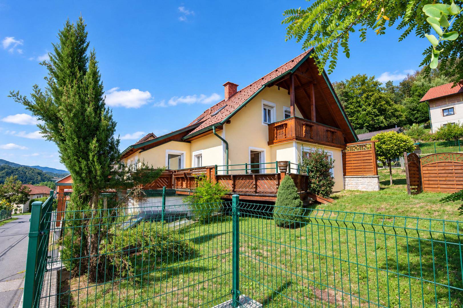 Gelbes Einfamilienhaus mit rotem Ziegeldach, umgeben von einem grünen Zaun und Bäumen.