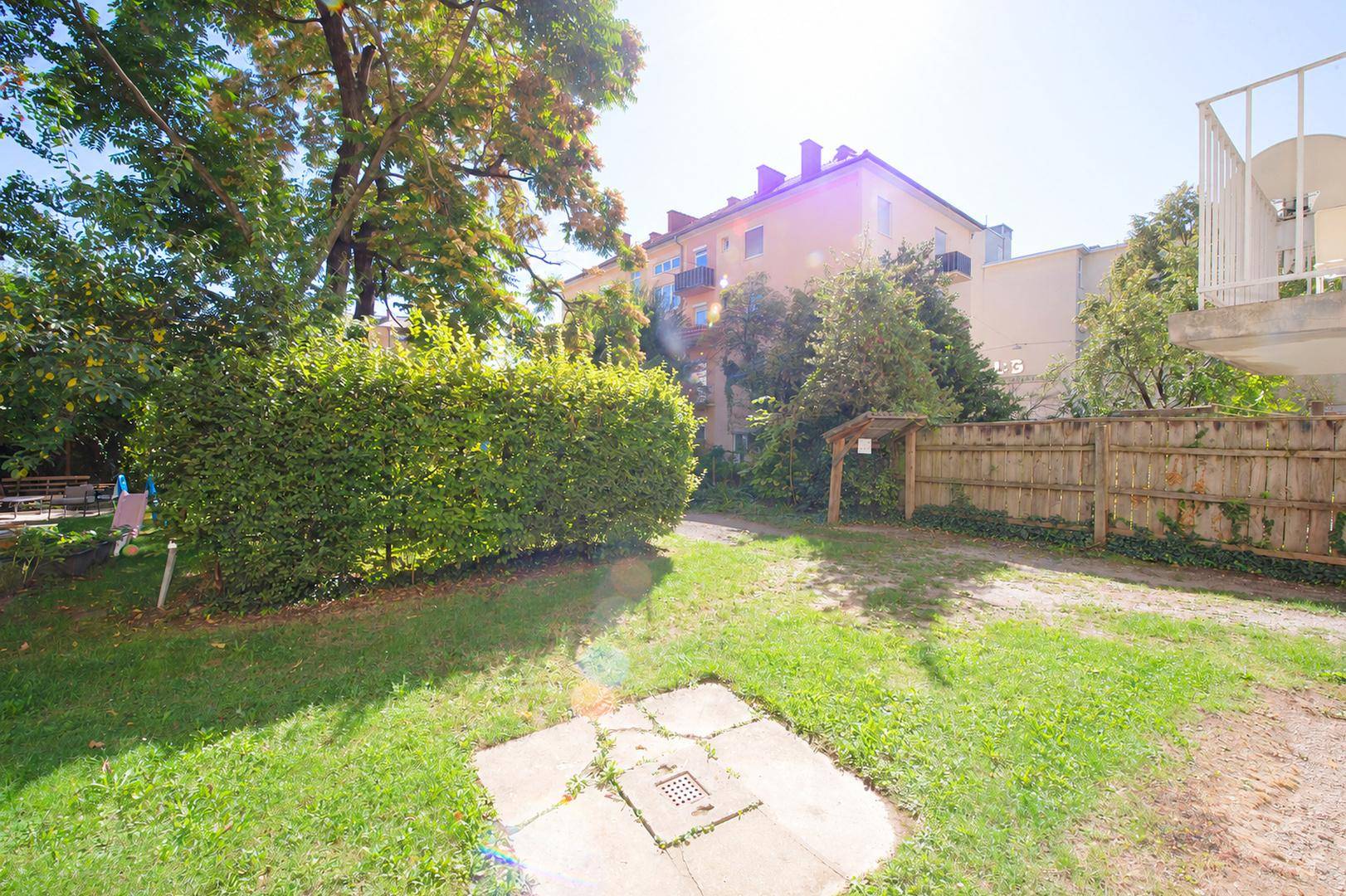 Sonniger Gartenbereich mit Rasenfläche, Hecke und einem Zaun, umgeben von Wohngebäuden.