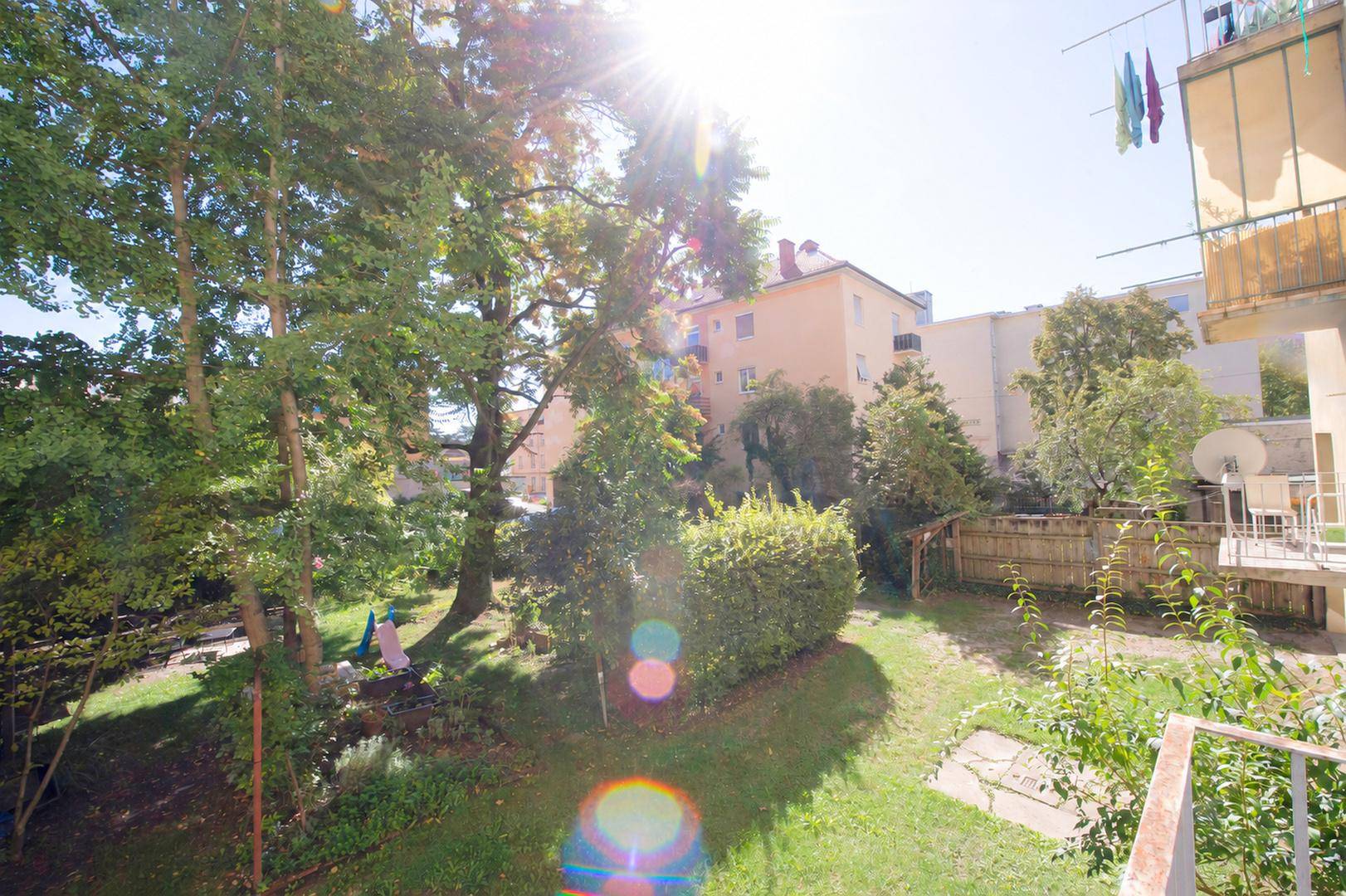 Sonniger Gartenbereich mit viel Grün, Bäumen und einem Ausblick auf die umliegenden Wohngebäude.