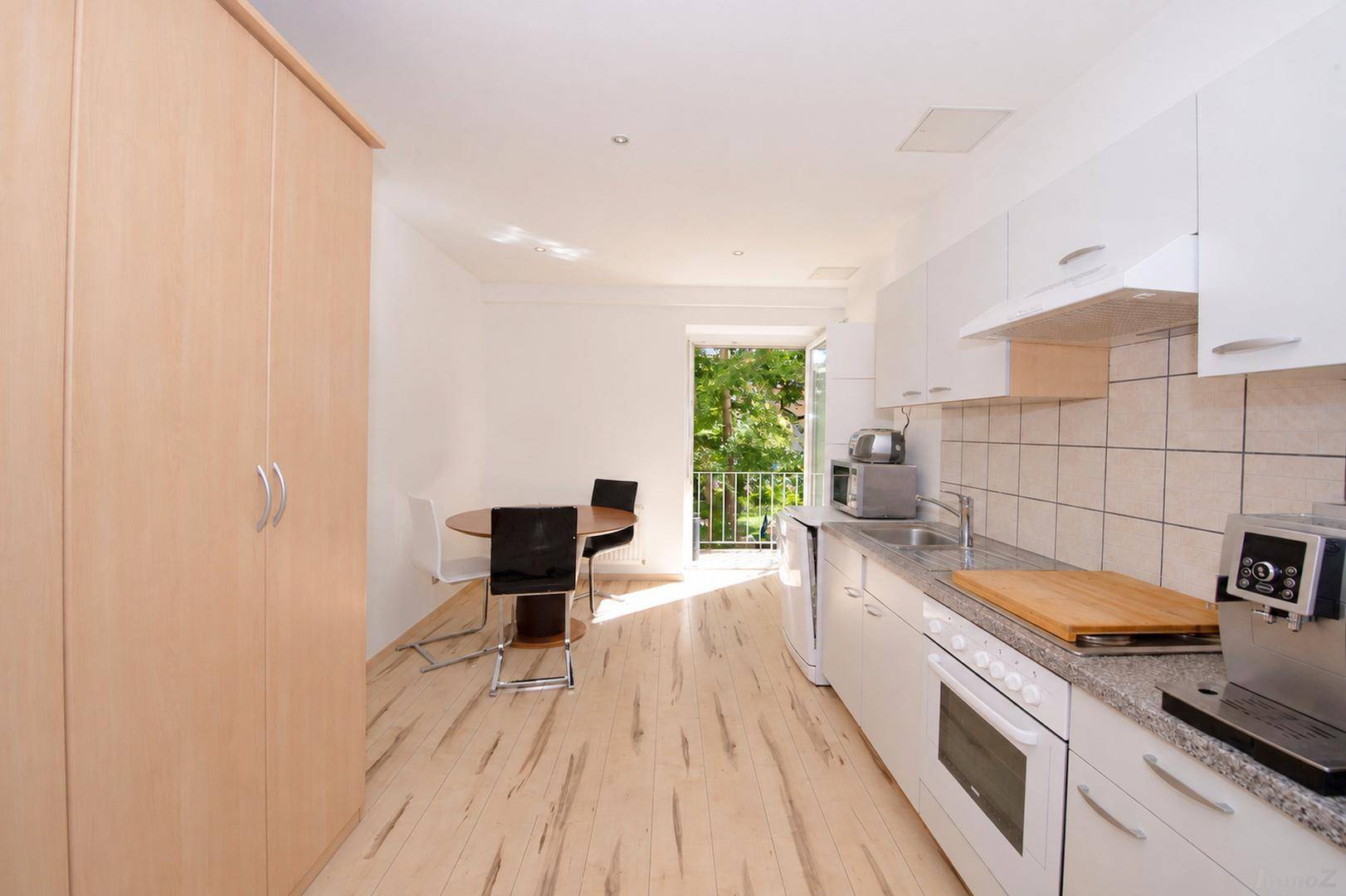 Helle Küche mit weißer Einbauküche, Esstisch und Zugang zu einem Balkon mit Grünblick.