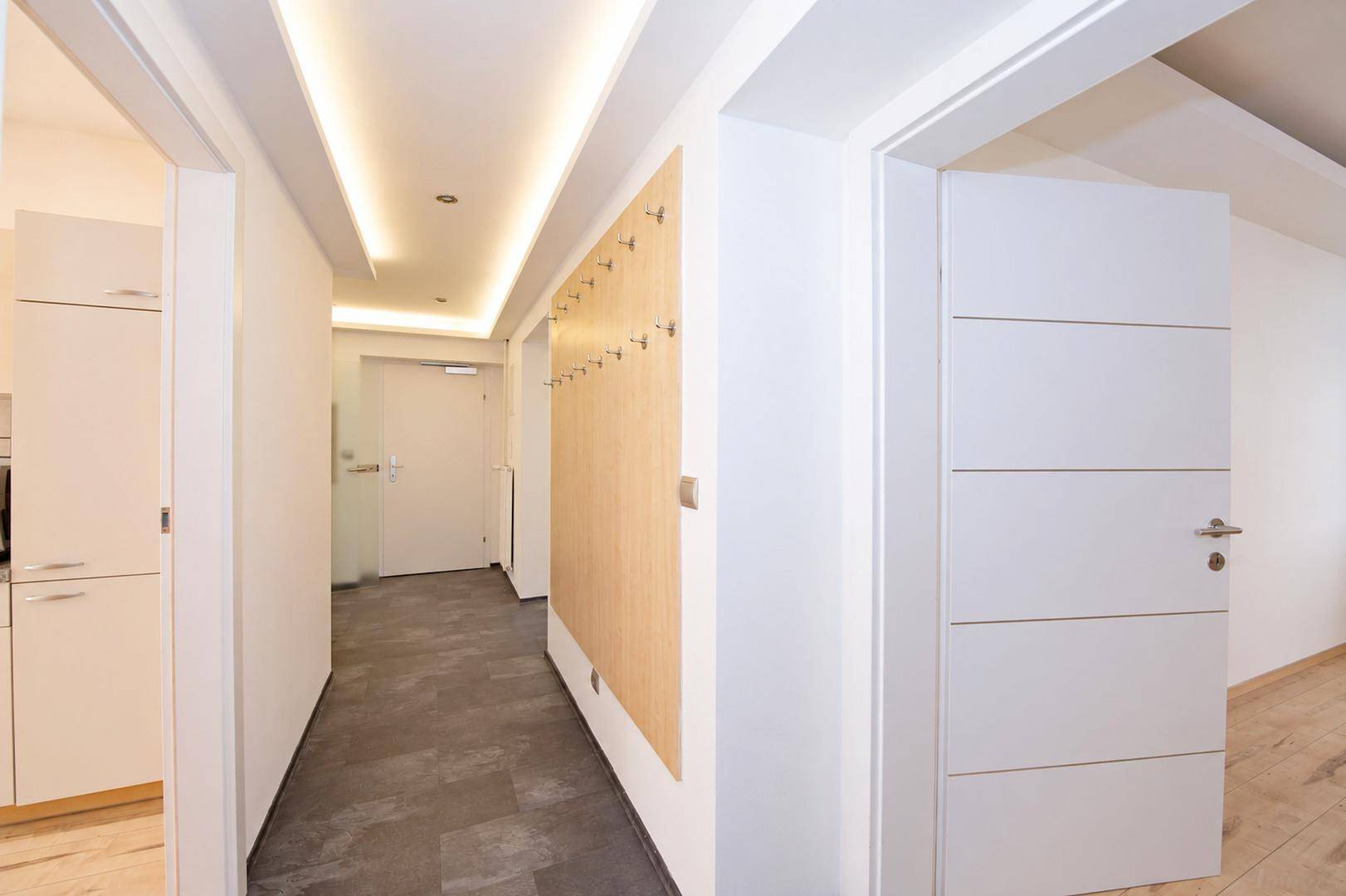 Heller Flur mit indirekter Beleuchtung, dunklem Fliesenboden und Garderobe, führt zur Küche.