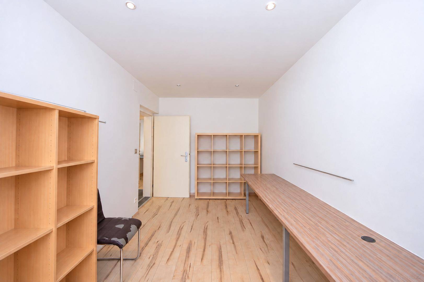 Büro mit hellem Holzboden, Regalen und langer Arbeitsplatte, Zugang zu einem weiteren Raum.