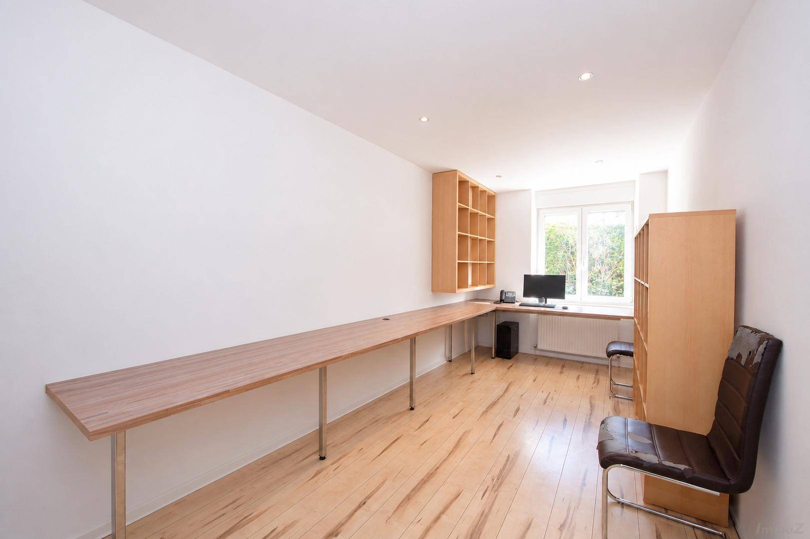 Langes Büro mit durchgehender Arbeitsplatte, Regalen und Fenster mit Blick ins Grüne.