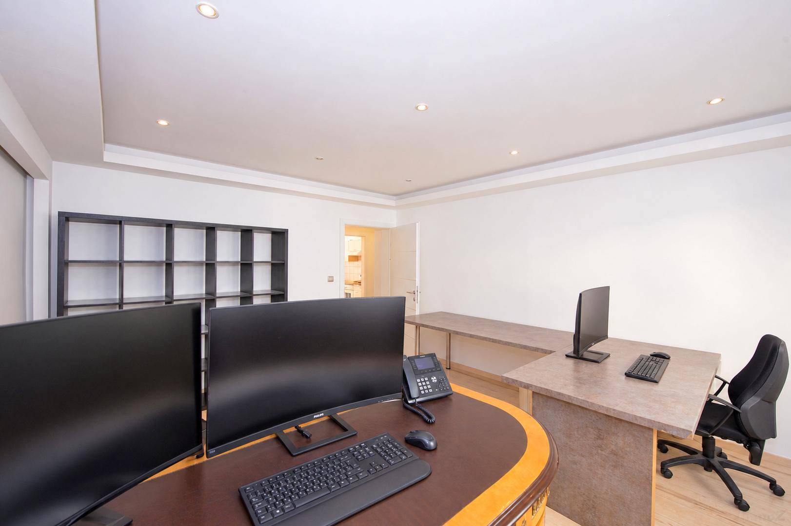 Büro mit mehreren Schreibtischen, Bürostühlen und einem dunklen Regal, heller Holzboden.